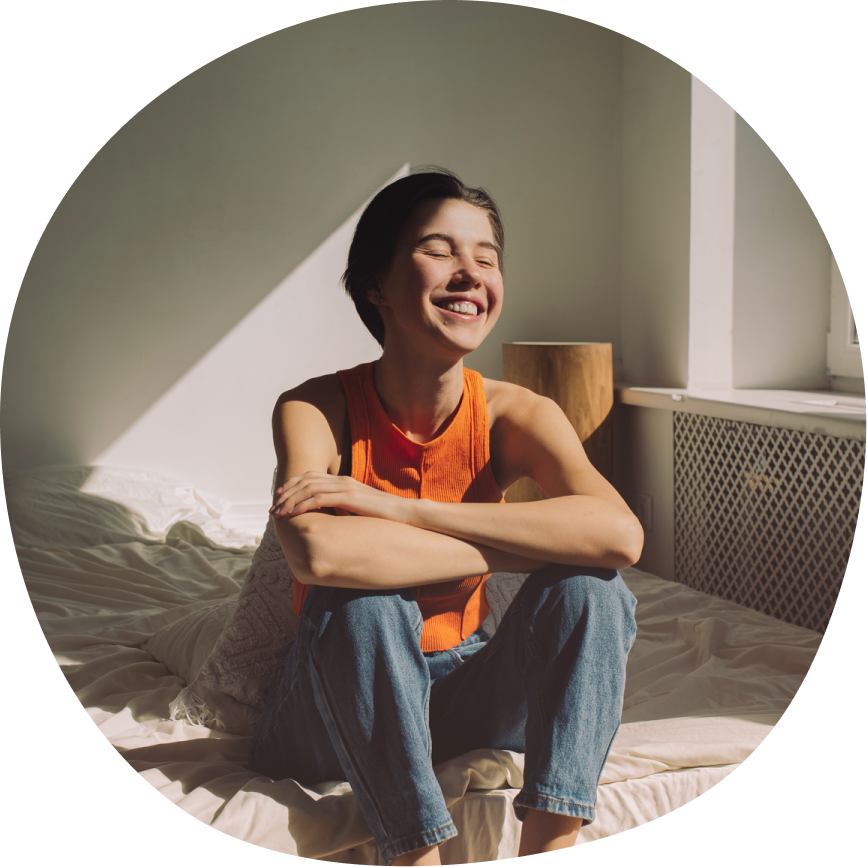 Young woman sitting cross-legged on bed with genuine smile and closed eyes, wearing orange tank top and jeans, bathed in natural sunlight